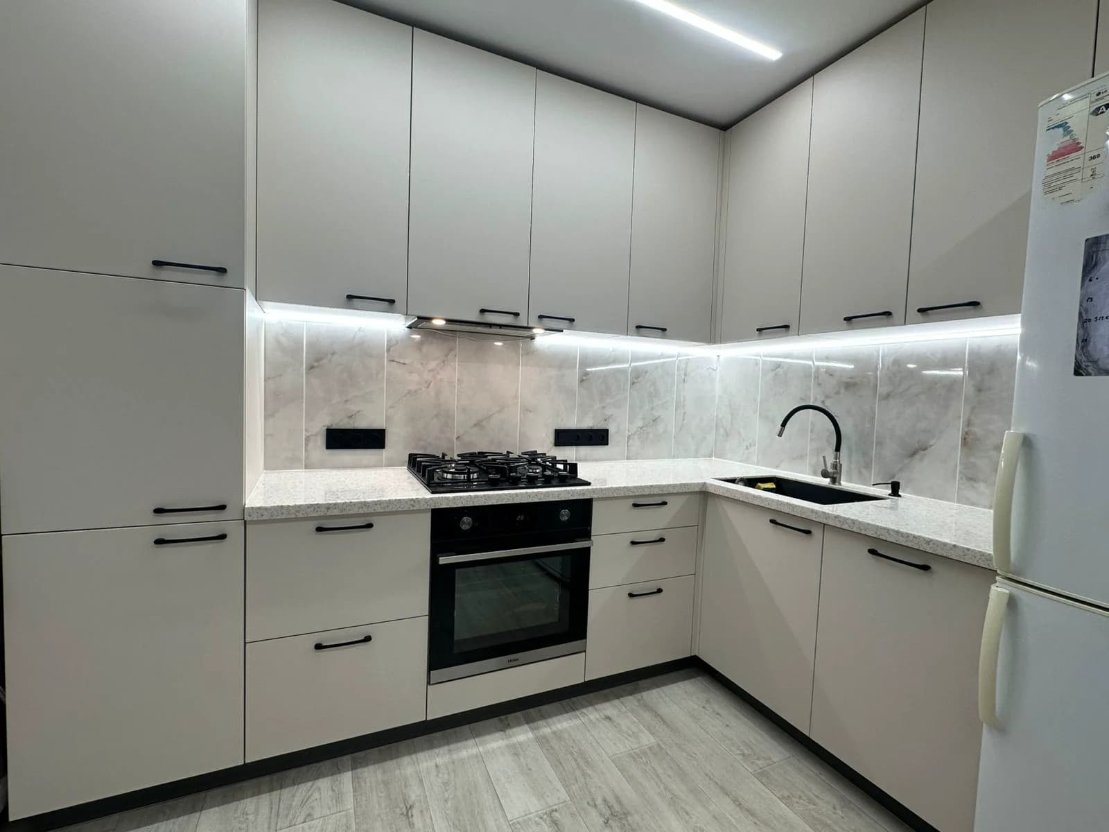 Белая угловая кухня с мраморным фартуком, чёрной фурнитурой и LED-подсветкой